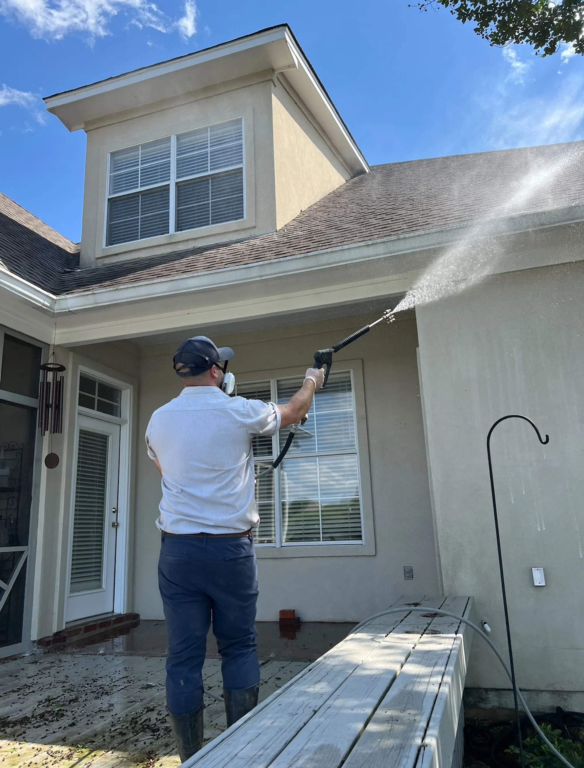 A uniformed technician soft-washing the upper siding and gutters of a stucco home with a long-reach sprayer