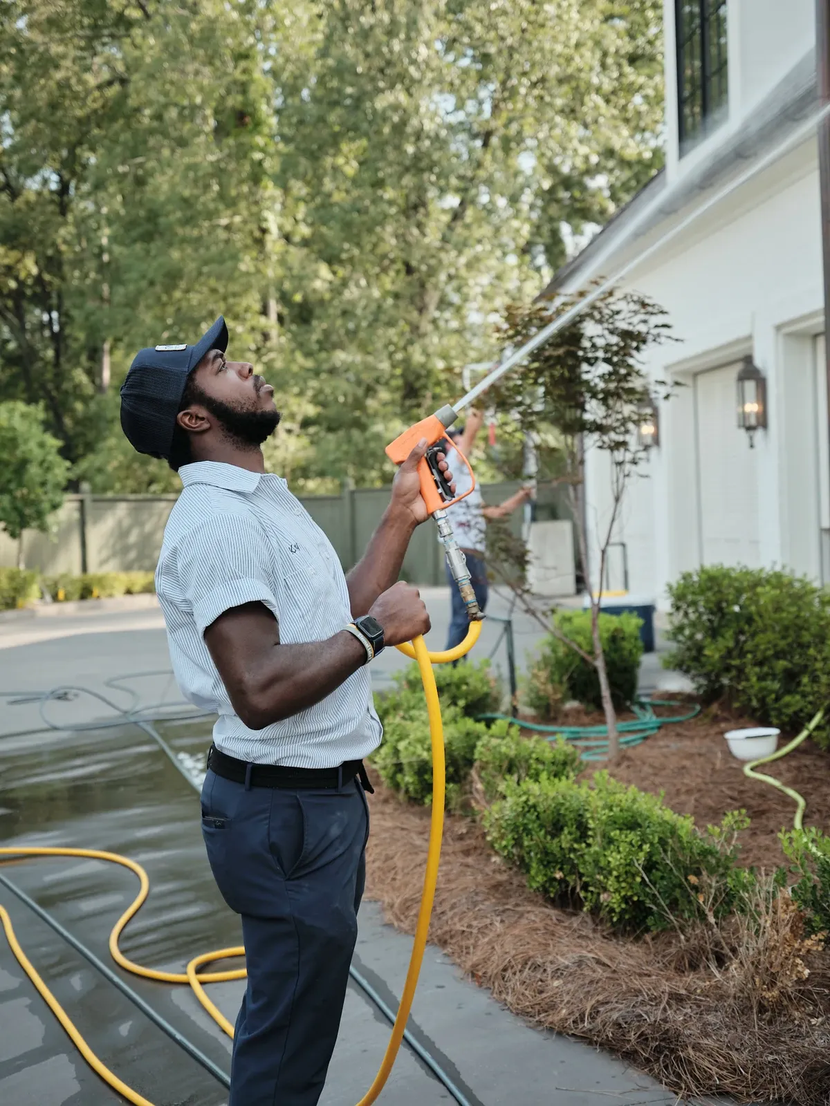 A uniformed exterior cleaning technician using a hose and sprayer to clean the side of a house